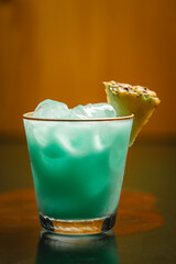 A blue cocktail with ice and a fresh pineapple slice on a table