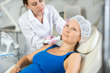 Young woman cosmetologist performs hardware facial ultrasound procedure to adult female patient