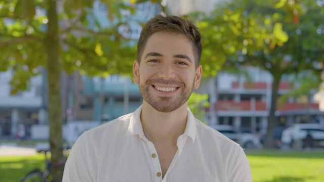 Handsome man looking at camera. Closeup positive guy smiling at camera. Portrait of male model posing on camera in green park. Cheerful man face with positive emotion outdoors
