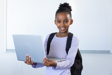 In school classroom, african american girl holding laptop and smiling with backpack