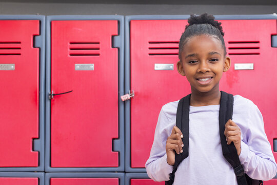 Smiling african american girl with backpack standing in front of red lockers at school, copy space
