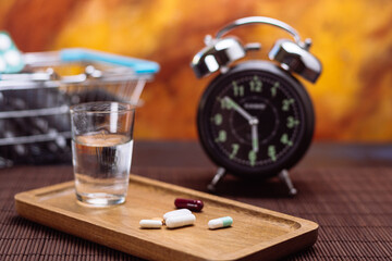 Variety of pills on wooden tray with water, alarm clock, and shopping basket