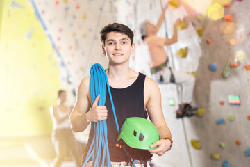 young positive muscular man with necessary equipment stands in gym near climbing wall. active...