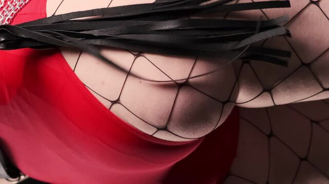 BDSM whip in the hands of a sexy girl dressed in a red bodysuit, a leather harness with a chain and a black background. Beats himself on the buttocks with a whip. BDSM - outfit for adult sex games
