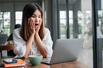 A young woman with a surprised expression looks at her laptop screen while sitting in a modern cafe with a cup of coffee.