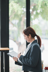 Professional woman in a modern office environment, writing on a digital tablet, with large windows and green outdoor scenery in the background.