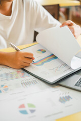 Close-up of a business professional analyzing financial charts and graphs on paper and laptop in an office setting.