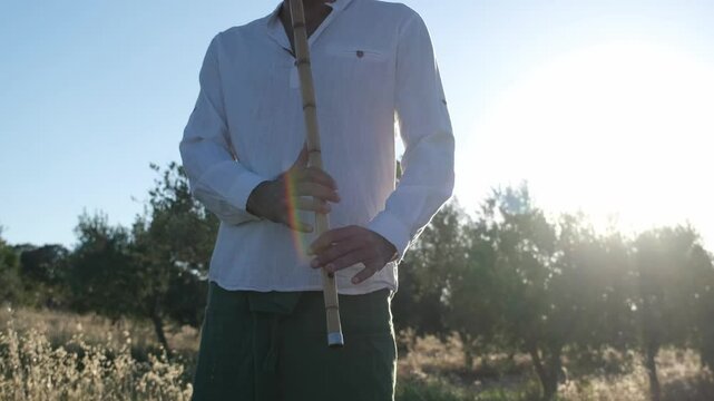 A Middle Eastern man in his 40s plays the ney in an olive grove during sunset, surrounded by nature and tall grasses, creating a peaceful and serene atmosphere