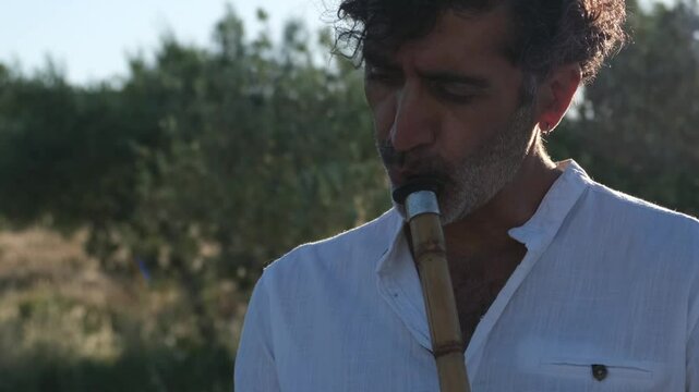 A Middle Eastern man in his 40s plays the ney in an olive grove during sunset, surrounded by nature and tall grasses, creating a peaceful and serene atmosphere
