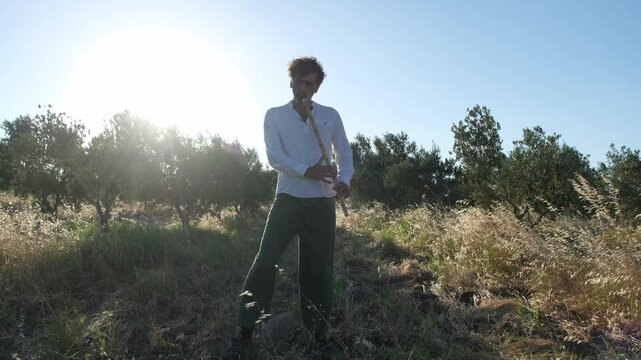 A Middle Eastern man in his 40s plays the ney in an olive grove during sunset, surrounded by nature and tall grasses, creating a peaceful and serene atmosphere