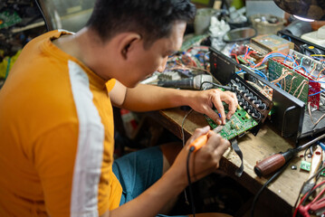 Asian repairman uses electric soldering iron to repair electronic circuit on workbench