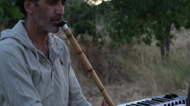 A Middle Eastern man in his 40s plays the ney in an olive grove during sunset, surrounded by nature and tall grasses, creating a peaceful and serene atmosphere
