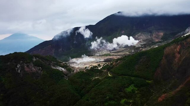 Aerial video of Mount Papandayan's volcanic crater, encircled by lush mountains under a sky with drifting clouds, capturing the dramatic and stunning beauty of this natural landscape in Indonesia.