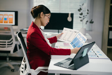Young asian woman is reviewing mobile application wireframes while working at her desk in the...