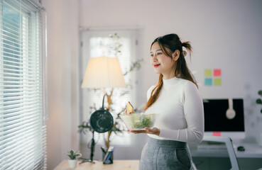 Young businesswoman is taking a break from work to eat a healthy salad in her home office. She is standing by the window and looking outside