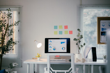 Modern, bright home office is pictured with a computer monitor displaying analytics charts and graphs. The desk is white with a white chair, keyboard, mouse, lamp, and potted plants