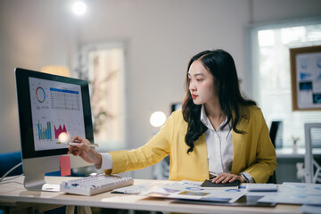 Young professional is reviewing charts and graphs on her computer screen, using a pen to point at the data.  She is surrounded by paperwork and working diligently at her desk