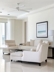 Coastal living room interior design featuring white furniture, a ceiling fan, and a large window with an ocean view. The room is decorated in a modern style with a coastal touch.