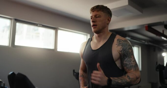 Confident and purposeful tattoo guy in a gray T-shirt runs on a treadmill during his warm-up before training in the fitness room in the morning