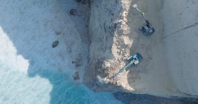 Top Down Drone Video of Turquoise Waves Crashing Against Environmental Destruction of the Limestone Cliffs near Pura Luhur Uluwatu Temple, Bali Indonesia with Excavators Dumping Earth into Ocean