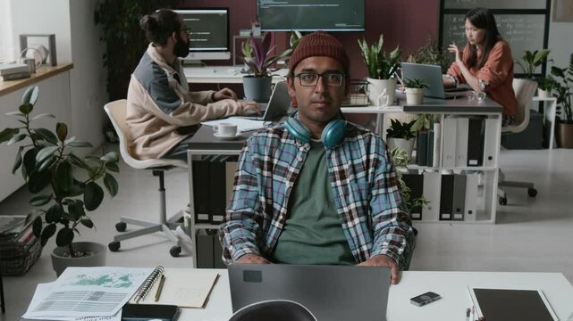Medium portrait of happy geeky Indian male software developer in checked shirt, beanie sitting with laptop at desk in office, headphones on neck, looking at camera, colleagues working in background