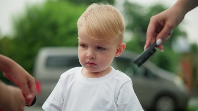 A young boy stares thoughtfully while a woman with painted nails gently combs his hair, the boy seems lost in thought, observing someone near him closely, a car is seen behind them