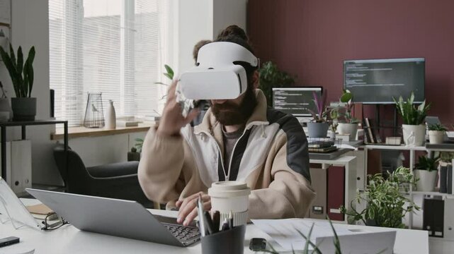 Medium full looped timelapse of young Middle Eastern male developer in AR headset working on virtual reality project at busy IT startup office during routine workday, writing code and testing