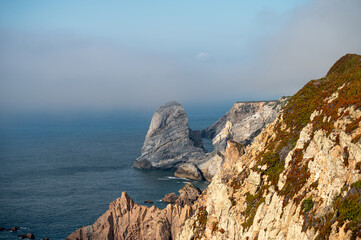 A rocky coastal cliff with mist surrounding the sea stacks in a serene afternoon light