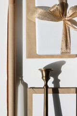 Christmas Festive Flatlay. Gift Box with Golden Ribbon Bow, Cards and Envelopes, Kraft Packaging Paper, Brass Candlestick, Candle on White Background. 