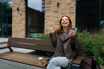 Cheerful redhead woman receiving unexpected good news in social media app using smartphone sitting on urban bench on summer day. Happy female celebrating success with joy and positive vibes.