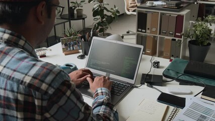 Medium close-up over-shoulder shot of male software engineer in checked shirt writing code on laptop, lines running on black screen, during routine workday in IT company office