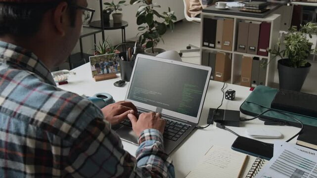 Medium close-up over-shoulder shot of male software engineer in checked shirt writing code on laptop, lines running on black screen, during routine workday in IT company office