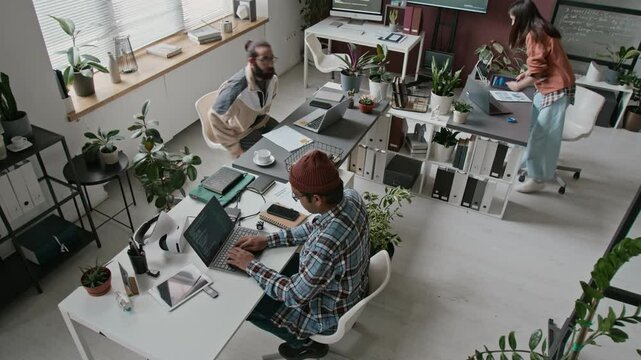 Wide top shot of three diverse, multiethnic male and female programmers working in open plan office at tech startup company - writing computer code, discussing projects, developing applications
