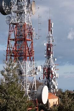 Two tall transmission towers with multiple antennas and satellite dishes stand amidst a natural environment, under a partly cloudy sky.