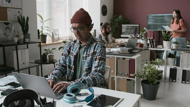 Medium full shot of geeky Indian male programmer in plaid shirt, beanie sitting at desk in IT startup office, typing code on laptop while working on AR mobile application