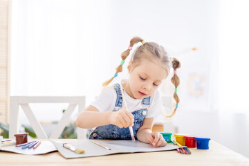 a little child girl paints a drawing with a brush in an album and smiles, a happy child draws a...
