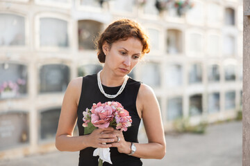 Sorrowful middle aged woman in black dress standing in cemetery holding bouquet of flowers with...