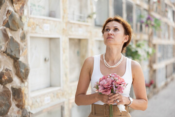 Elegant grieving woman in forties, wearing white tank top and pearls, standing with bouquet in...