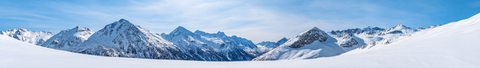 Obraz premium This image captures a wide panorama of snow-covered mountains, stretching across the horizon. The towering peaks are bathed in soft, natural light, creating a serene, wintry scene. The undulating