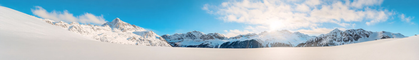 A stunning panorama of a snow-covered mountain landscape, where the rugged peaks dominate the horizon. The soft, powdery snow contrasts beautifully with the deep blue sky, while the distant peaks