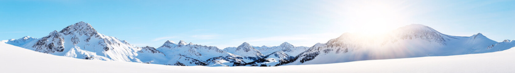 A wide panoramic shot of a snow-covered mountain range under a bright, clear sky. The jagged mountain peaks are blanketed in thick snow, their edges sharply defined against the vibrant backdrop. The