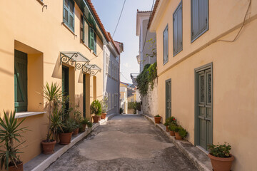 A peaceful alley in Nafplio features charming pastel buildings adorned with green shutters, surrounded by lush potted plants bathed in sunlight, inviting exploration.