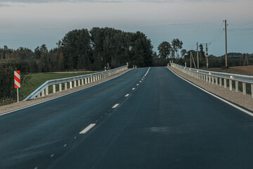 Fototapeta premium A smooth, empty road winds through a wooded area with dense trees on one side and open fields on the other. The soft evening light creates a peaceful atmosphere with ample copy space in the sky and on
