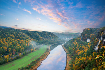 Fototapeta premium Scenic view of the Elbe River winding through colorful autumn forests in Saxon Switzerland National Park, Germany. Misty valley under a dramatic sunrise sky