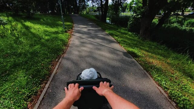 A point-of-view shot of a person pushing a stroller along a paved path in a green park. The stroller holds a child wearing a hat. The scene is calm, sunny, and surrounded by nature.