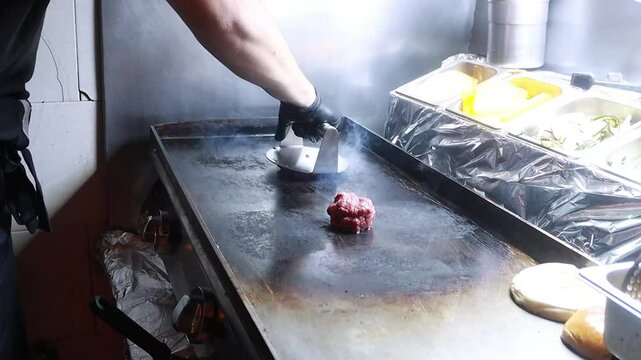 Video of a unrecognizable person pressing raw smash burger balls in a hot griddle in a fast food restaurant, flattening them to cook in a professional kitchen