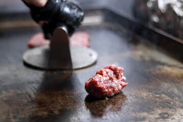 Gloved hand flattening a raw smash burger ball on a hot griddle, searing it into a burger in a...