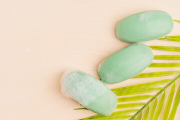 Top view of three wet hygienic craft antibacterial soap bars of green colour soapy and covered with foamy bubbles on beige yellow background with copy space and palm leaf used as toiletries for body