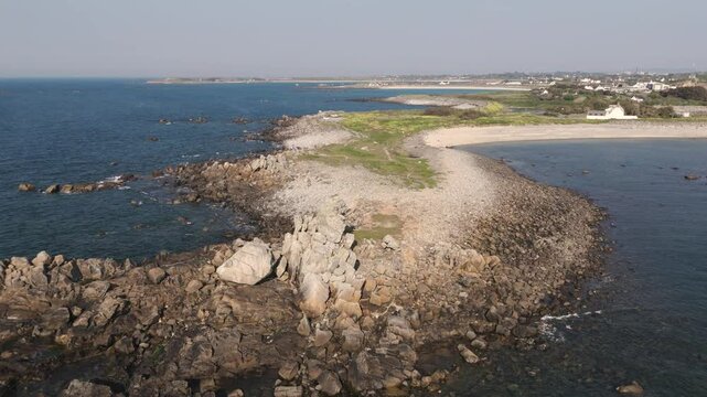 Flight over rocky outcrop towards golden beach and distant bays in Guernsey on bright sunny day