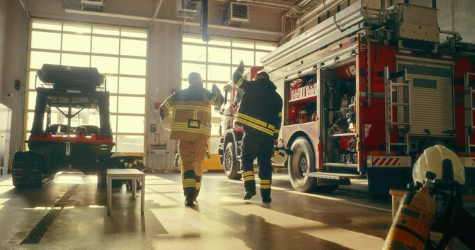 In the Fire Station Garage, Two Firefighters Walk Together Next to a Fire Truck. They Raise Their Hands for a High Five, Celebrating Their Efforts. Warm Color Grade Slow Motion Footage